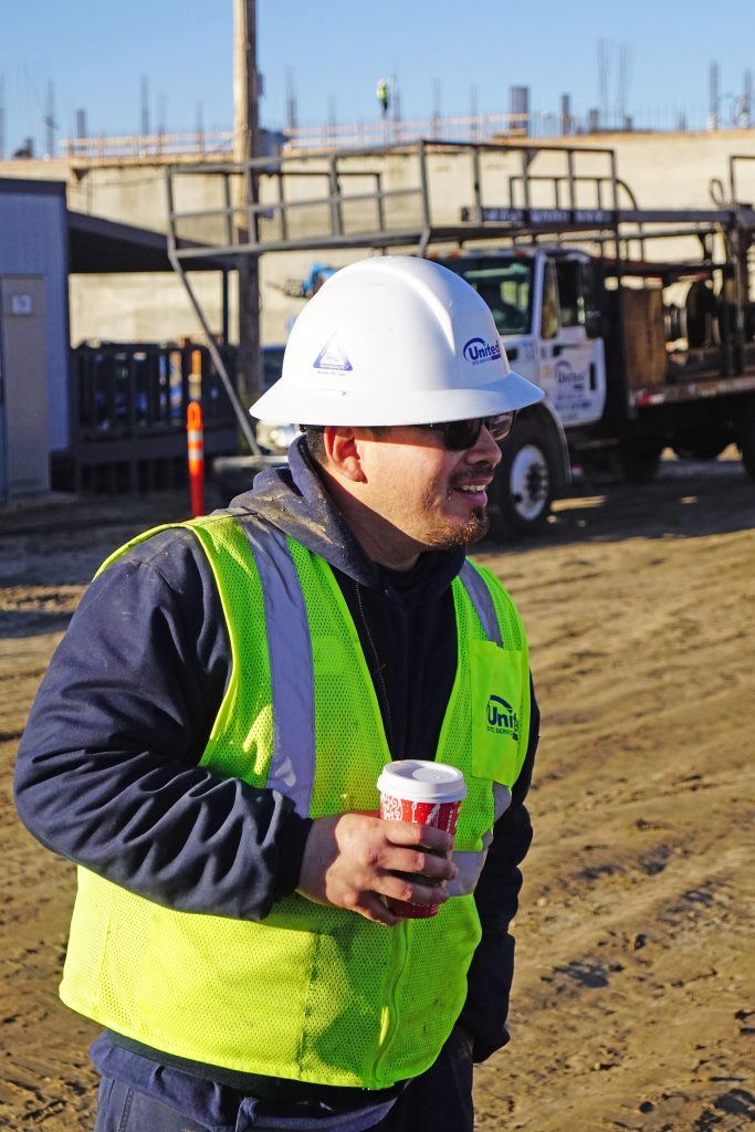 A construction worker wearing a high visibility jacket, hard hat, and safety glasses holds a cup of coffee on a construction site, smiling as he looks to the side