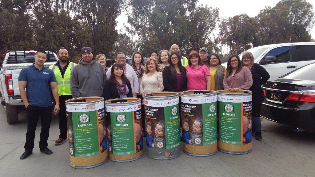 A group of staff smiling and standing behind three large donation barrels during the food drive