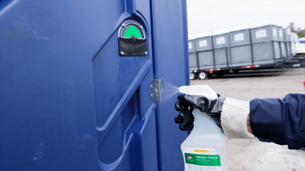 A person wearing a glove sanitizing the handle of a blue portable toilet with a spray bottle, with multiple trailers carrying several porta potties in the background