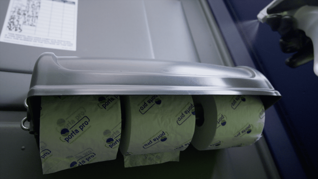 A gloved hand spraying sanitizer on the toilet paper dispenser in a portable restroom