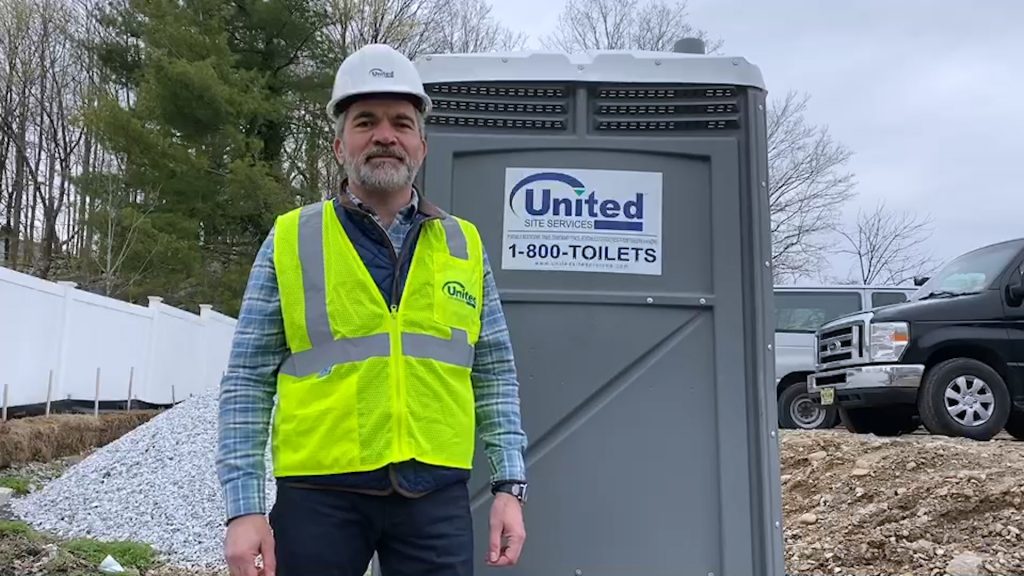 A man wearing a hard hat and hi-vis vest stands in front of a portable toilet outside