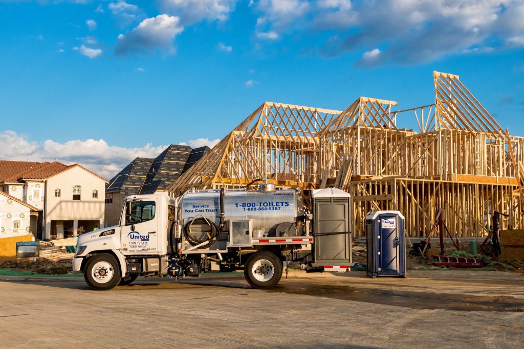 A construction site featuring a wooden house frame under construction, with a clear blue sky. A sanitation truck labeled "United Site Services" and portable toilets are parked nearby