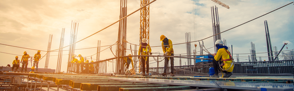 Construction workers in safety gear working together on a building site with metal beams and a clear sky at sunset