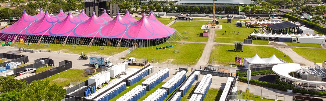 Aerial view of an outdoor event with multiple pink peaked tents and rows of white temporary structures on a sunny day, surrounded by greenery