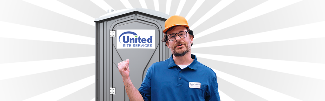 A worker in a blue uniform, glasses and hard hat standing in front of a portable toilet smiling and pointing backward at the unit