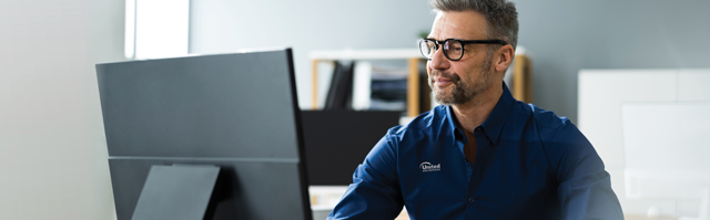 A male united site services employee wearing glasses and a blue shirt with a badge working intently at a computer in a modern office