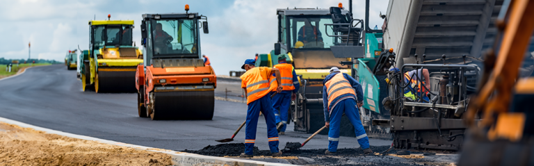 Road construction workers in high visibility jackets using tools to lay asphalt, with machinery like a steamroller and paver around them on a partly cloudy day