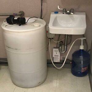 An ambient pump system sink consisting of a large white plastic barrel connected by hoses to a sink and a blue water jug