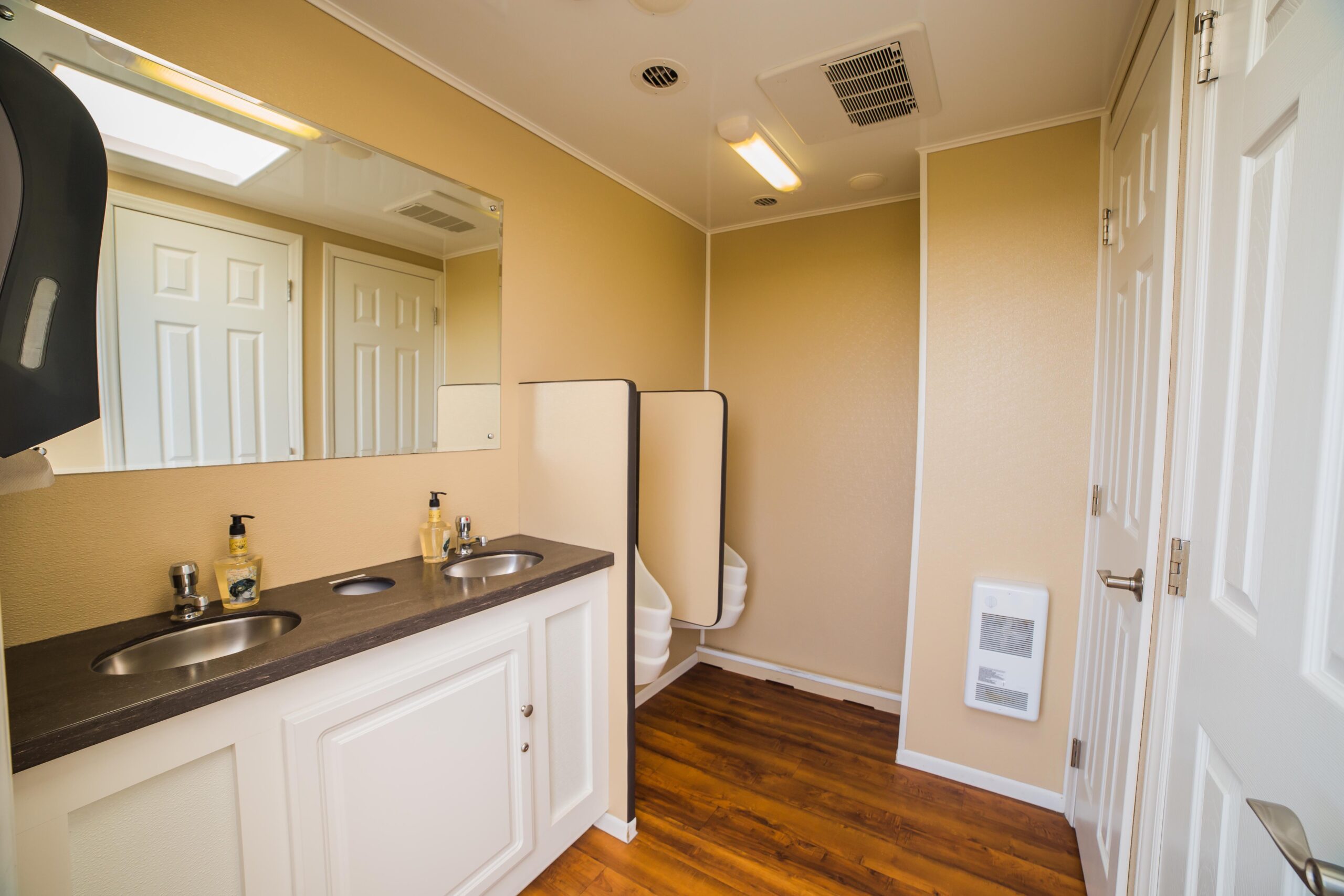 A clean trailer restroom with two sinks, mirrors, a partitioned area with urinals, and wooden flooring