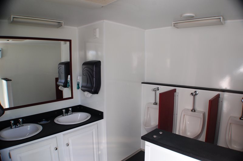 Interior of a clean portable restroom trailer with two sinks, a large mirror, soap dispensers, and three urinals against a white wall