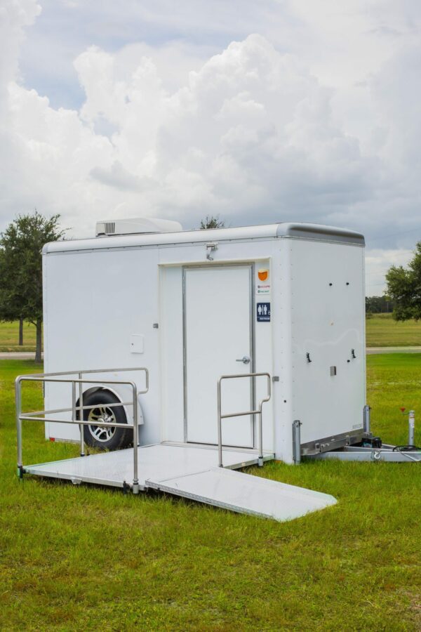 A white mobile trailer with a ramp and handrails parked on a green grassy field under a cloudy sky