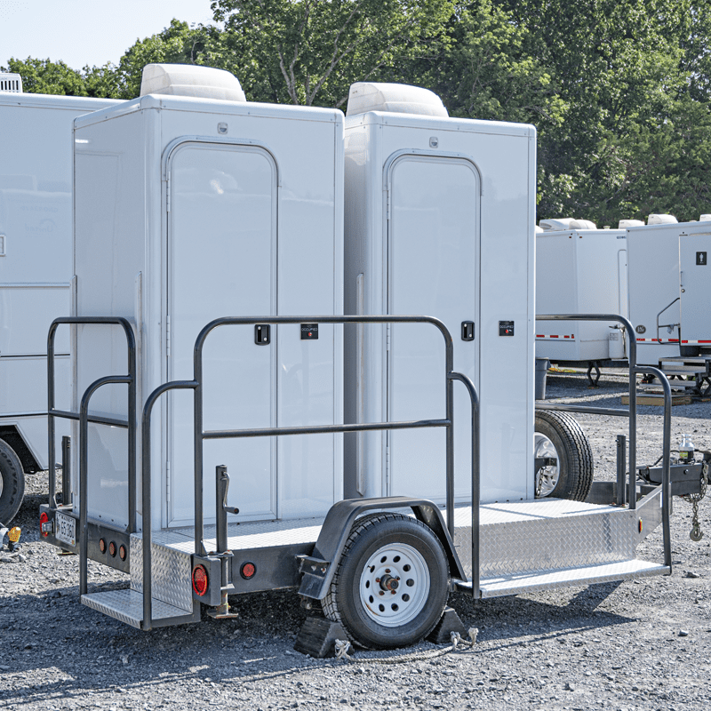 Two mobile solar-powered restroom trailers parked on a gravel lot, equipped with steps and railings, and each trailer connected to a small towing wheel assembly