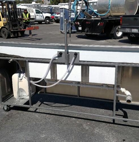 Back view of a portable 3-compartment sink with a 3-gallon water heater and a 250-gallon waste tank, located in a busy outdoor area with other utility vehicles.