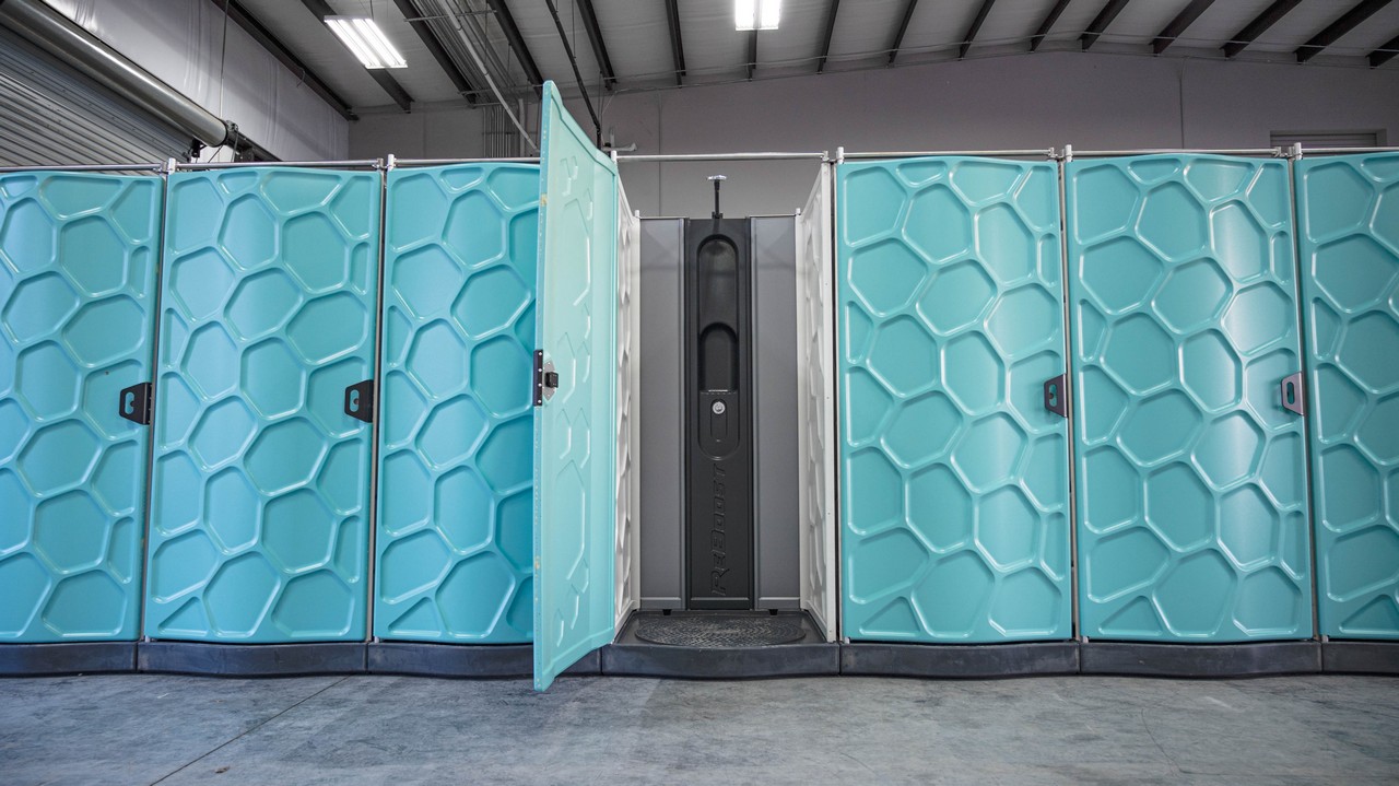 A row of turquoise Hydroflow portable showers with a door to one stall open inside a warehouse facility